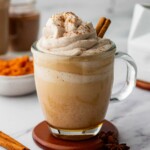 A clear glass mug filled with a pumpkin spice latte topped with whipped cream and sprinkled with cinnamon. Two cinnamon sticks garnish the drink. A saucer, cinnamon sticks, and star anise are visible nearby.