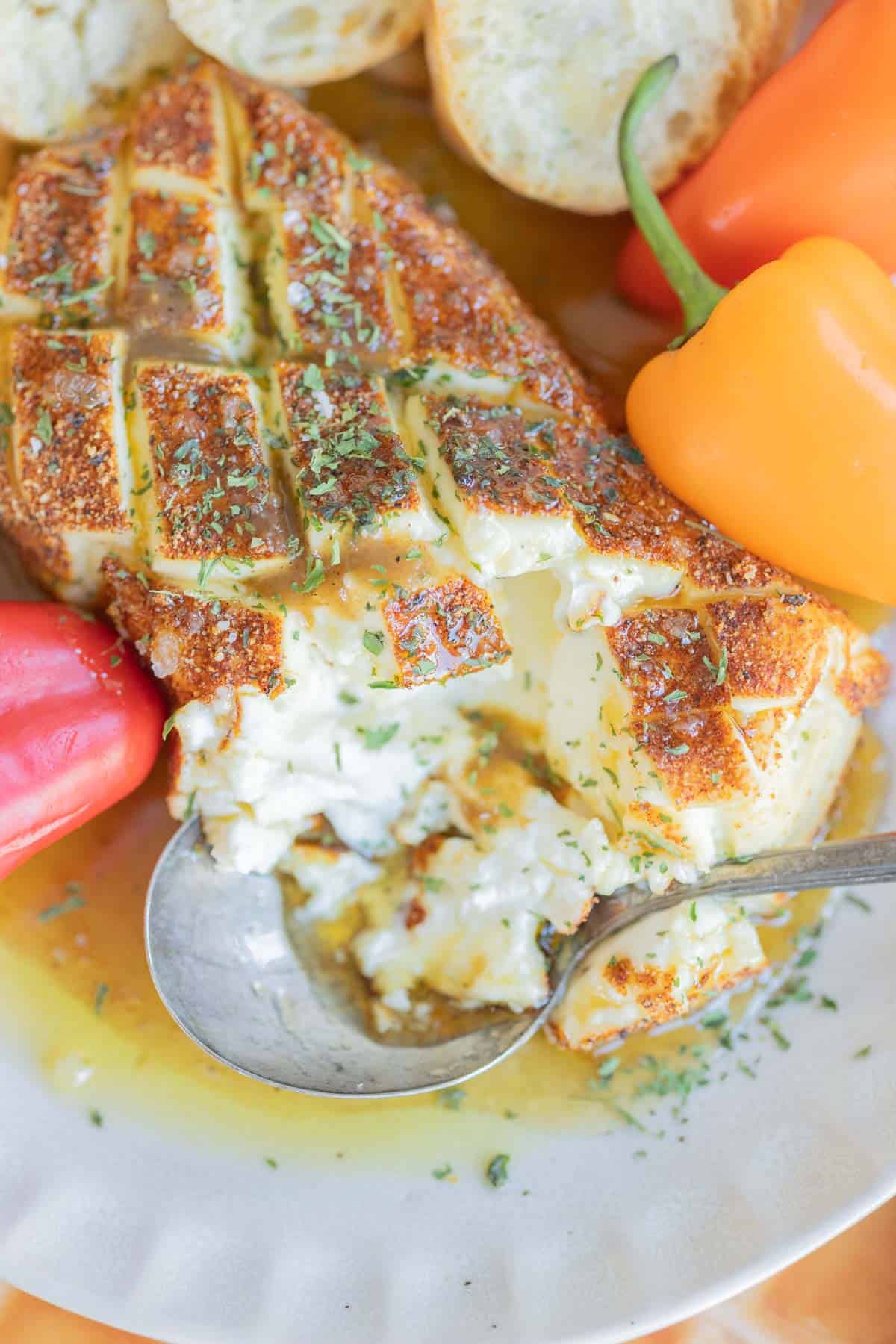 Smoked cream cheese with a golden, seasoned crust, garnished with herbs, surrounded by sliced bread and peppers, with a spoon scooping some cheese.