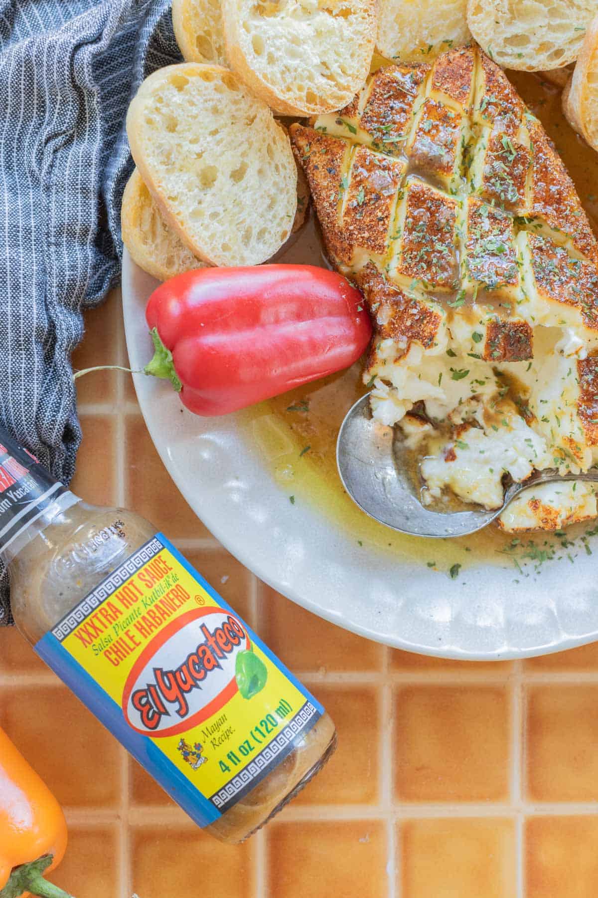 Grilled bread with smoked cream cheese, red pepper, and El Yucateco sauce on a plate, accompanied by sliced baguette on a tiled surface.
