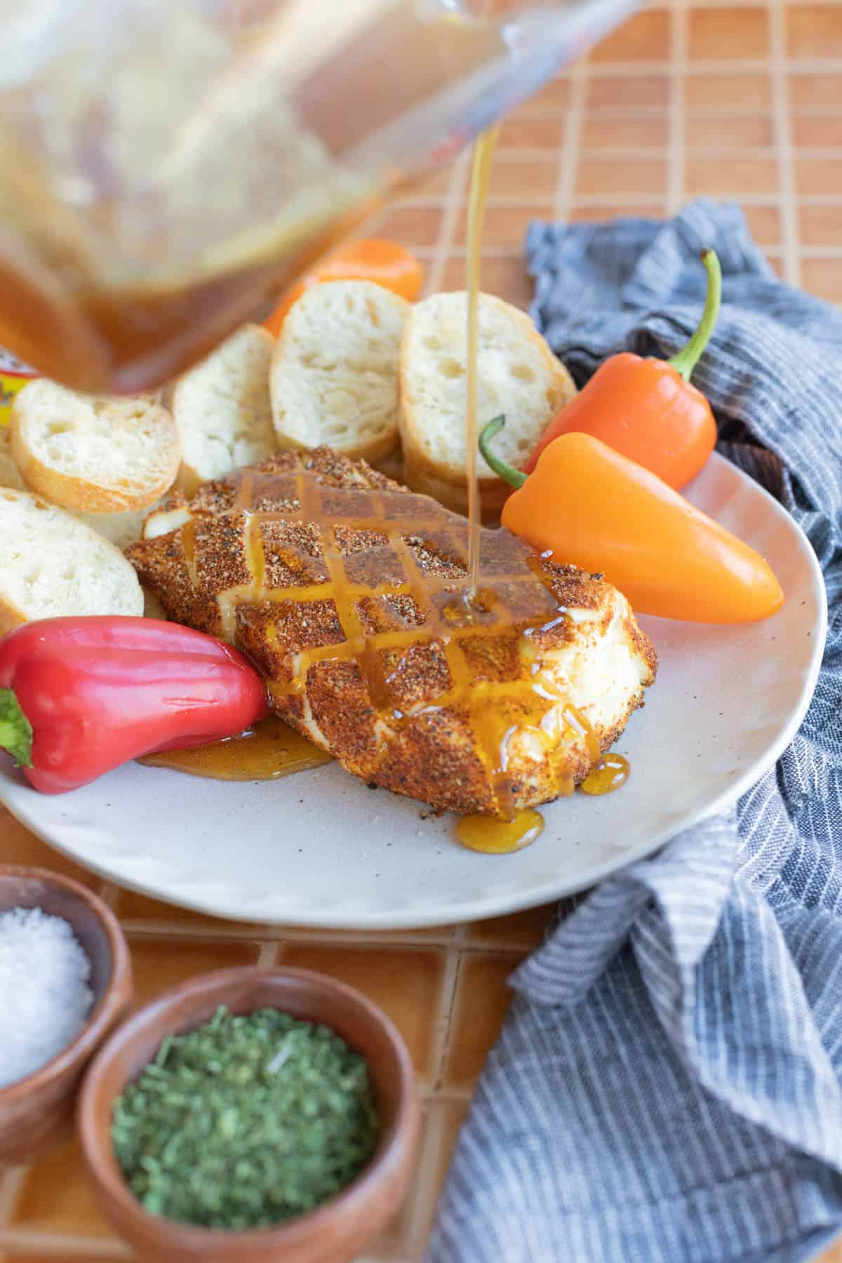 A plate with smoked cream cheese topped with sauce, surrounded by sliced bread and bell peppers. Small bowls with herbs and salt are nearby on a tiled surface.