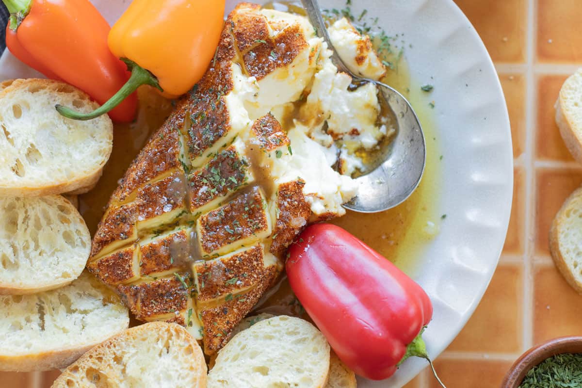 A plate with baked ricotta drizzled with honey and herbs, surrounded by sliced baguette and mini bell peppers.