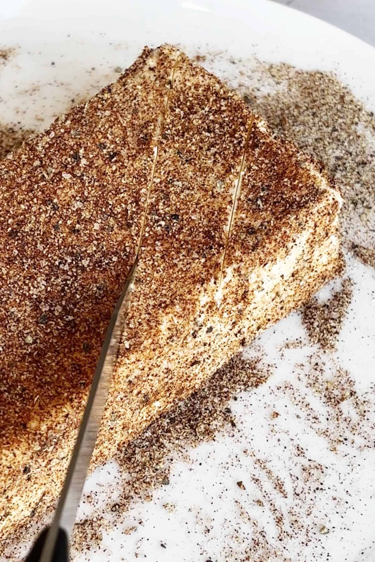 A knife slices through a spice-coated rectangular block of smoked cream cheese on a white plate.
