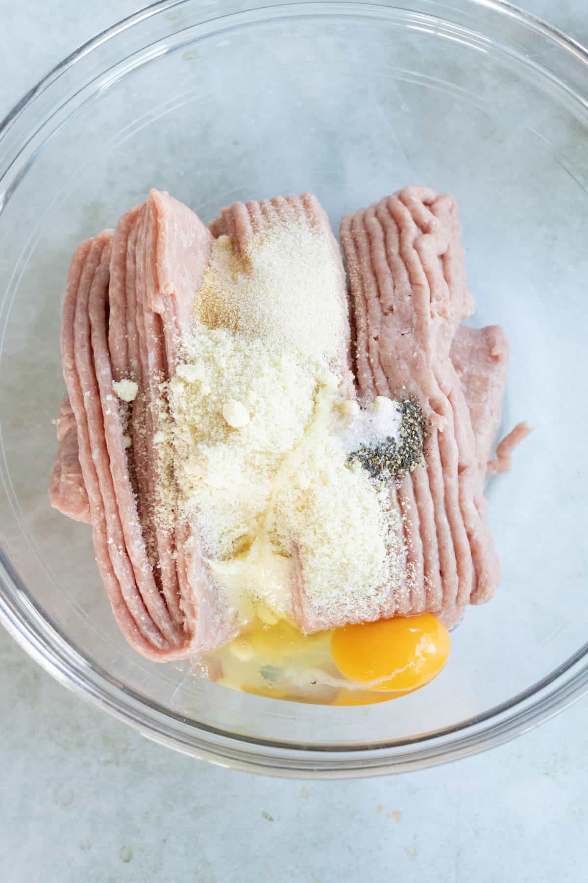 Ground turkey with an egg, breadcrumbs, and seasoning in a clear bowl on a light surface.