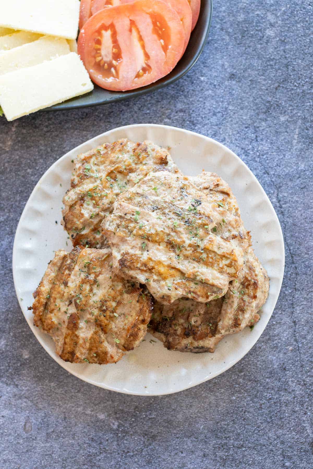 A plate of grilled turkey burgers on a gray plate, with sliced tomatoes and cheese nearby.