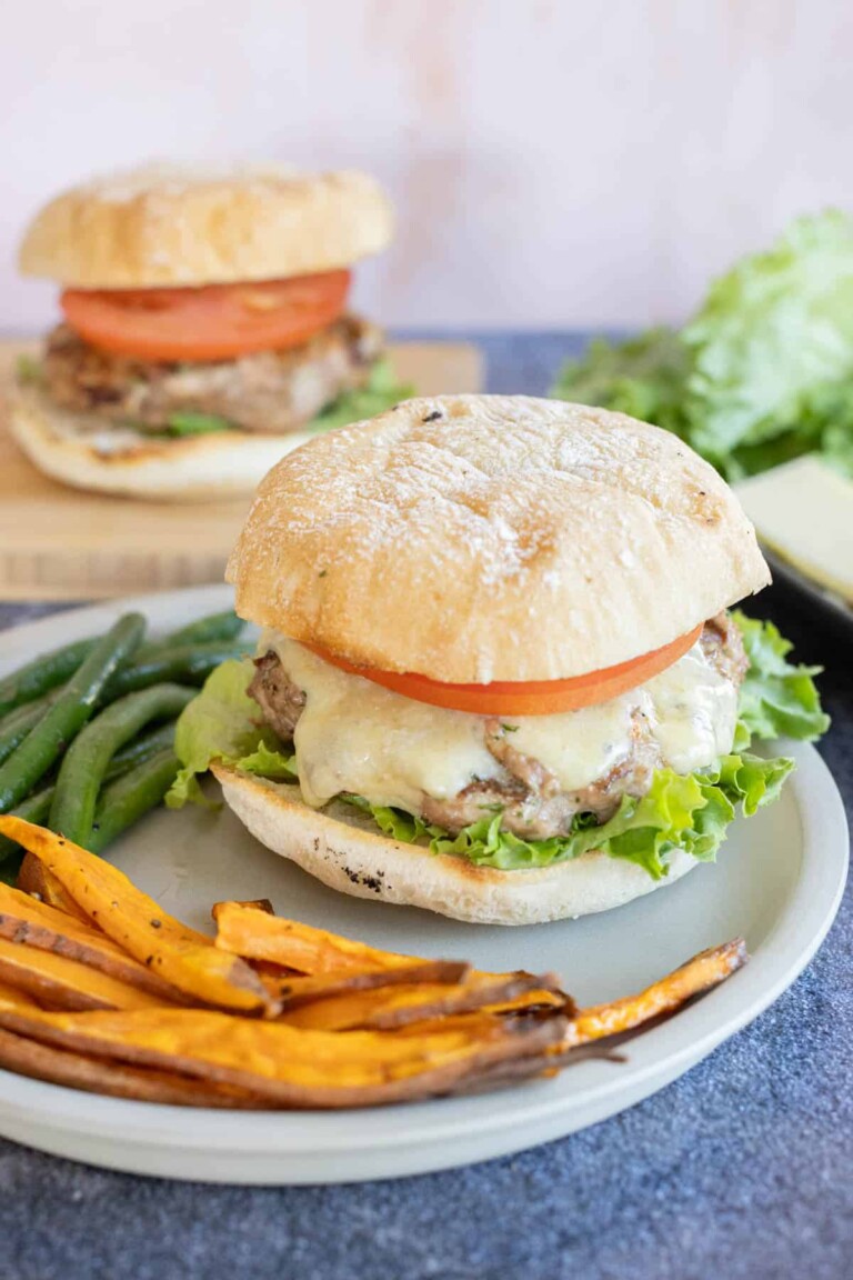 A turkey burger with melted cheese, lettuce and tomato on a plate, accompanied by sweet potato fries and green beans. Another burger is in the background.