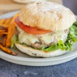 A turkey burger with lettuce, a patty topped with melted cheese, and a tomato slice on ciabatta bread, served with sweet potato fries on a plate.