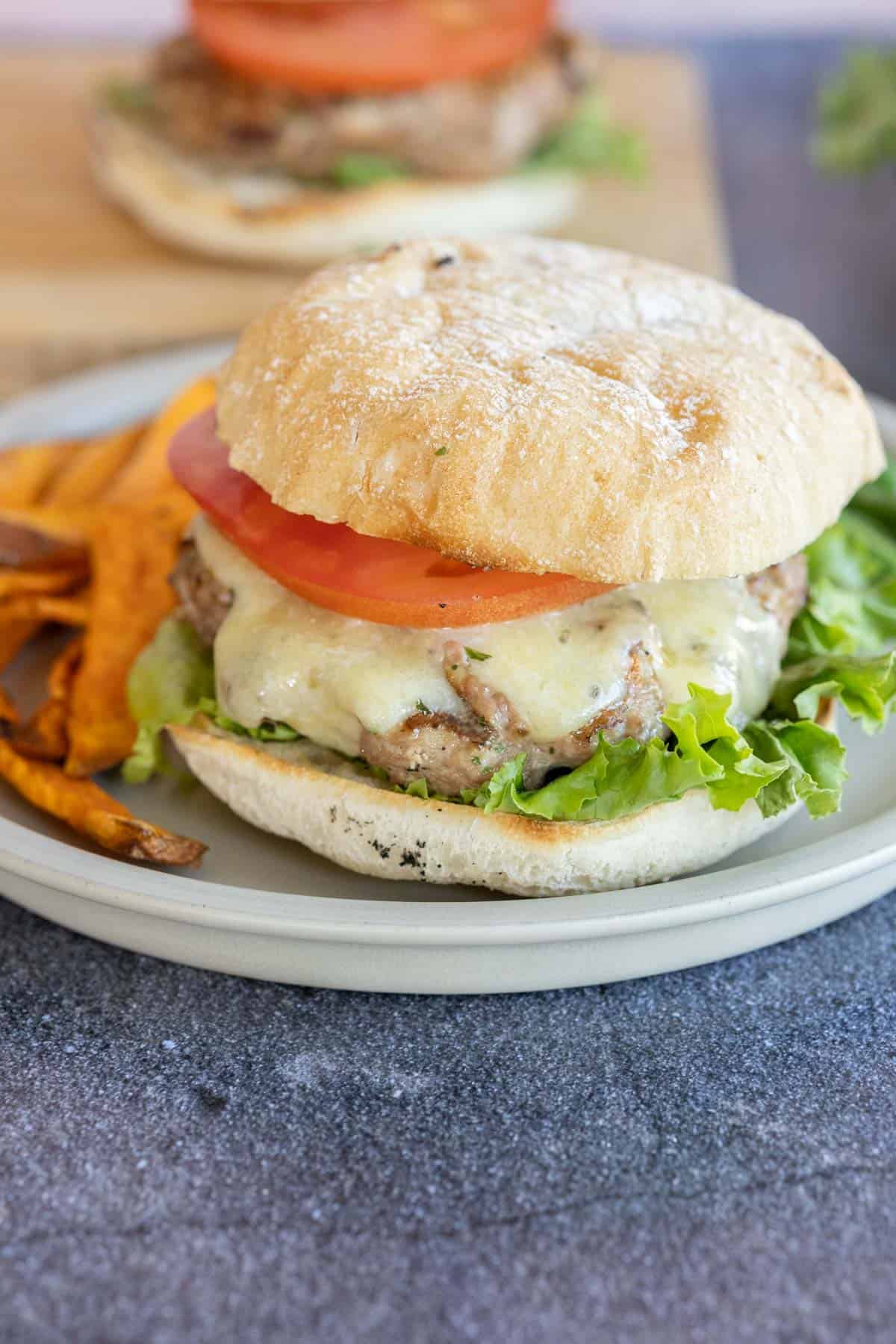 A turkey burger with lettuce, a patty topped with melted cheese, and a tomato slice on ciabatta bread, served with sweet potato fries on a plate.