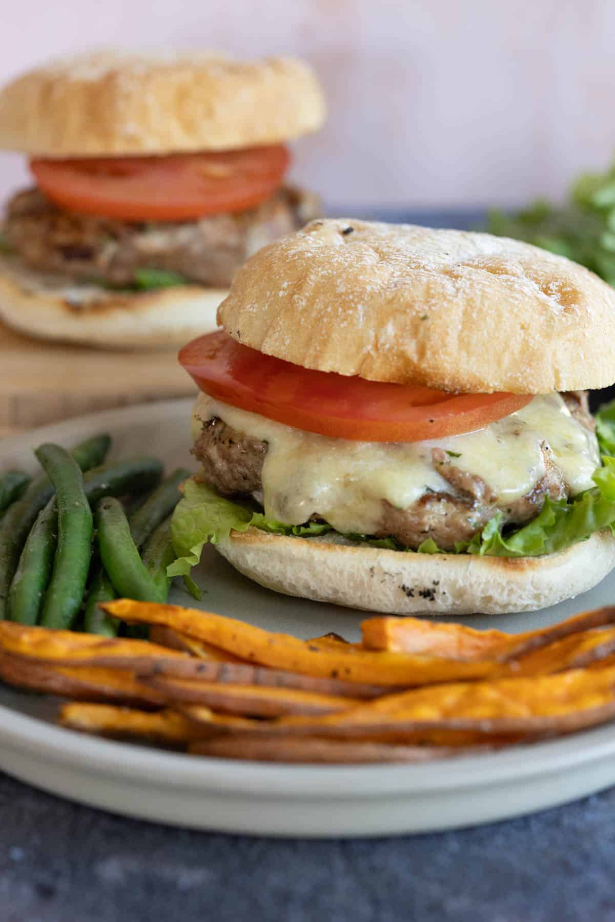 Two turkey burgers with lettuce, tomato, and cheese on buns, served with green beans and sweet potato fries on a plate.