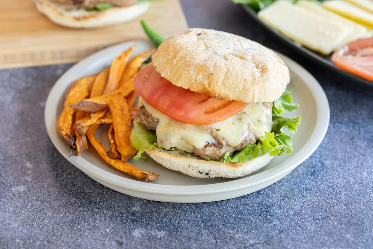 A turkey burger with cheese, lettuce and tomato on a ciabatta bun, served with a side of sweet potato fries on a round plate.