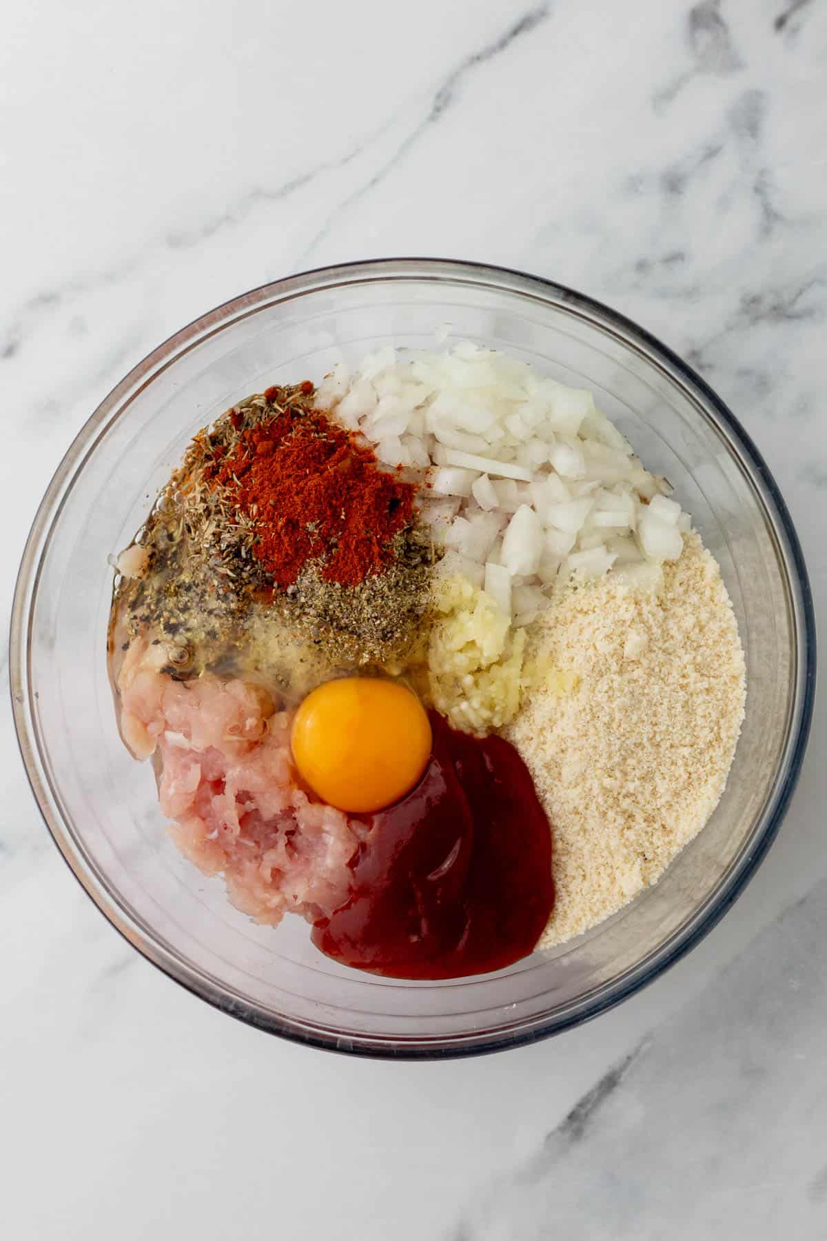 Bowl with raw ground turkey, chopped onions, egg, almond flour, ketchup, and spices on a marble surface.