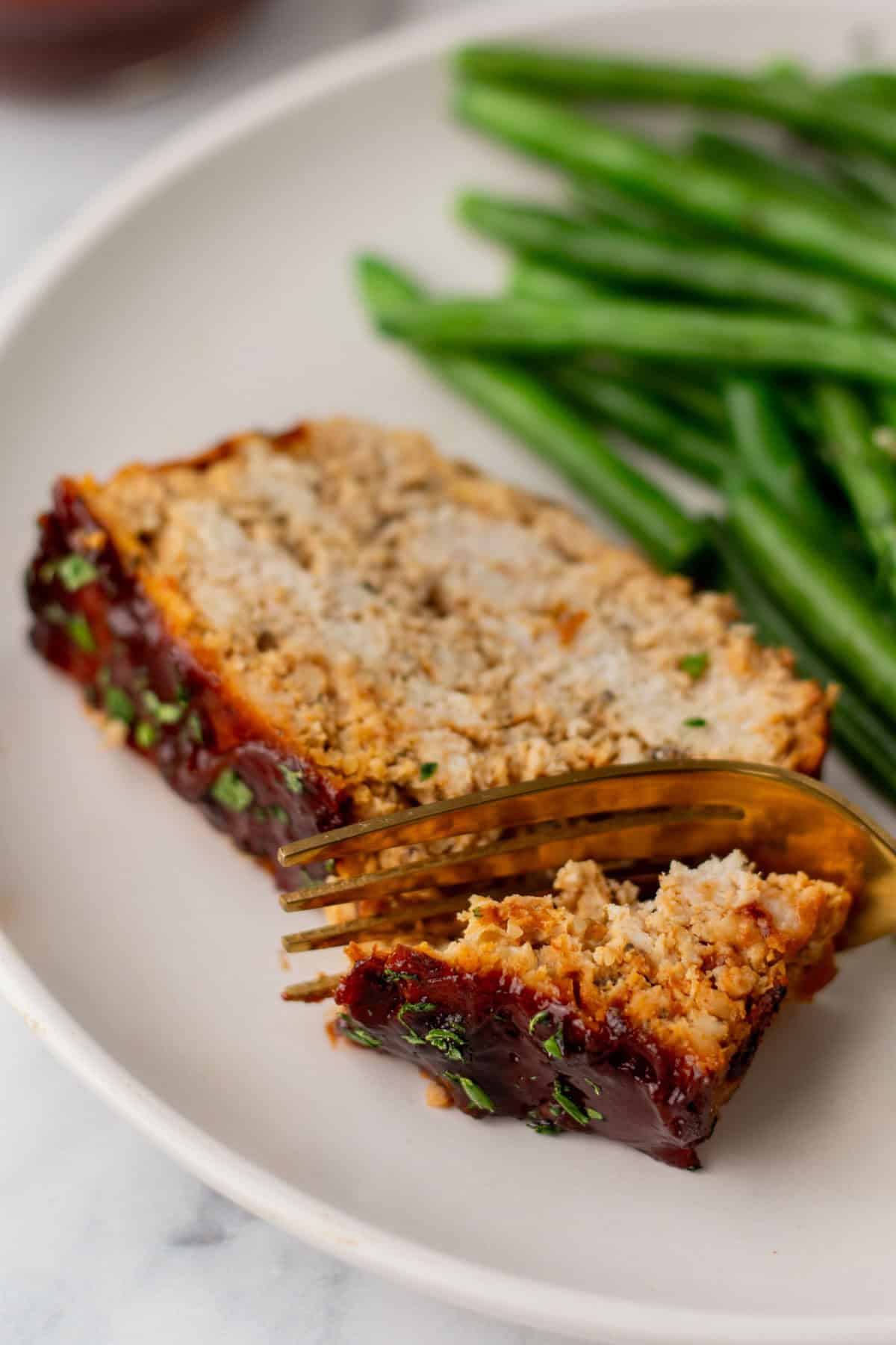 A slice of turkey meatloaf with a fork on a plate, accompanied by green beans.