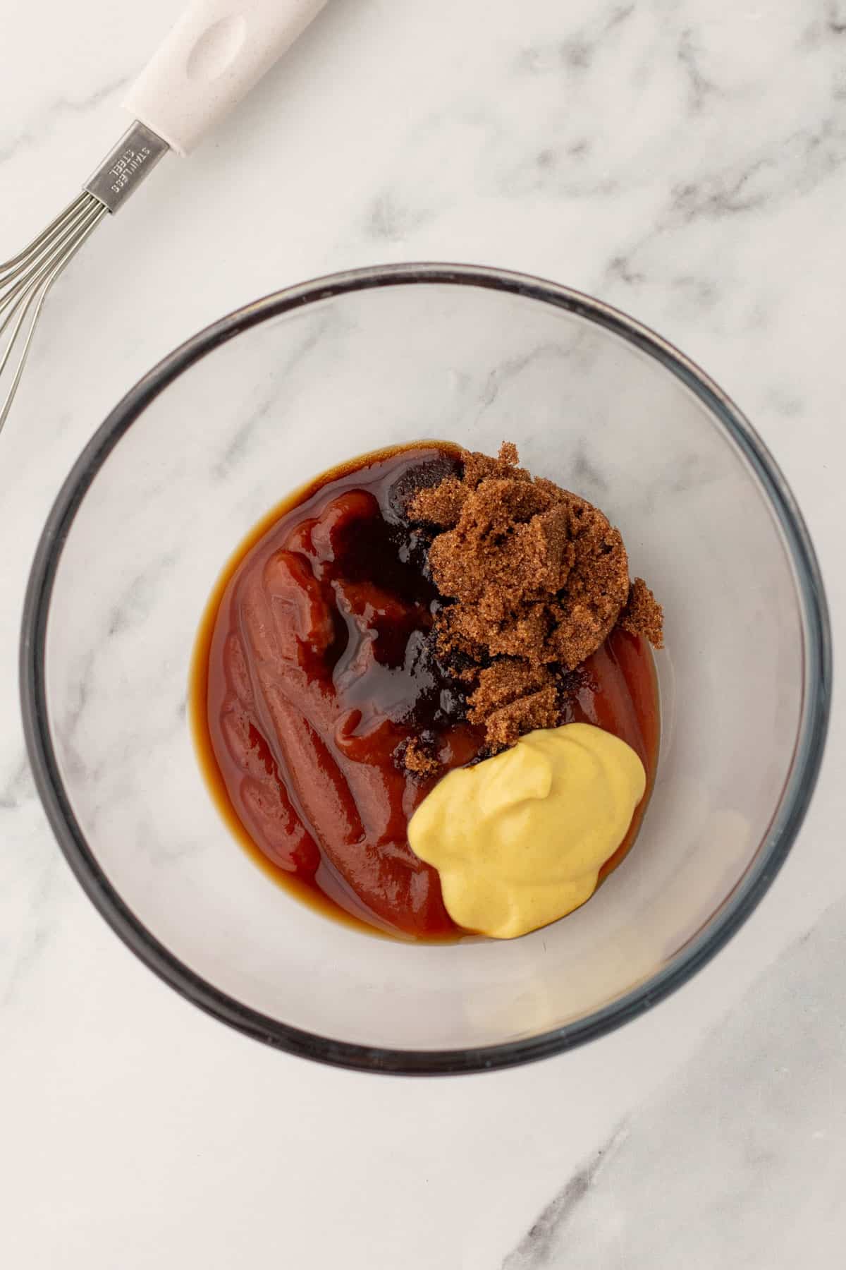 A glass bowl contains ketchup, mustard, soy sauce, and brown sugar on a marble surface.