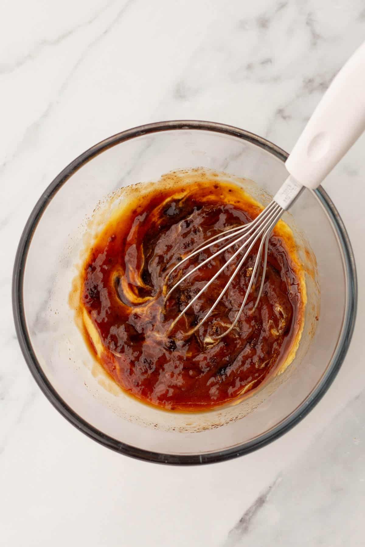 A glass bowl contains a whisk and a mixed brown sauce with visible spices, placed on a marble surface.