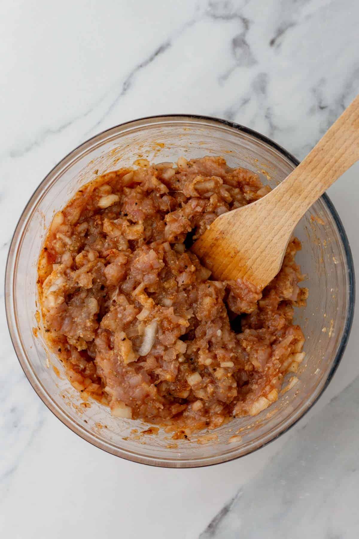 A glass bowl containing a mixture of ground turkey, chopped onions, and spices being stirred with a wooden spoon, on a marble countertop.