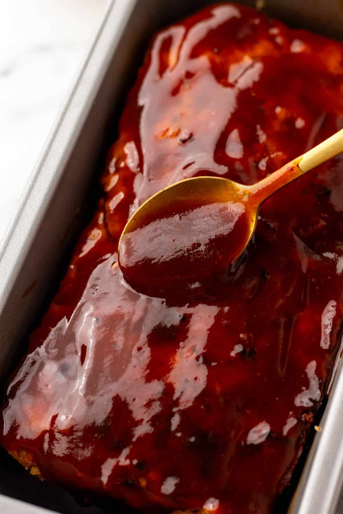 A spoon spreading red sauce over a meatloaf in a baking pan.