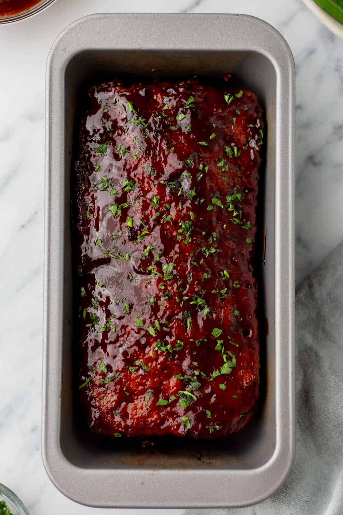 A glazed turkey meatloaf in a baking pan, garnished with chopped herbs, on a marble surface.