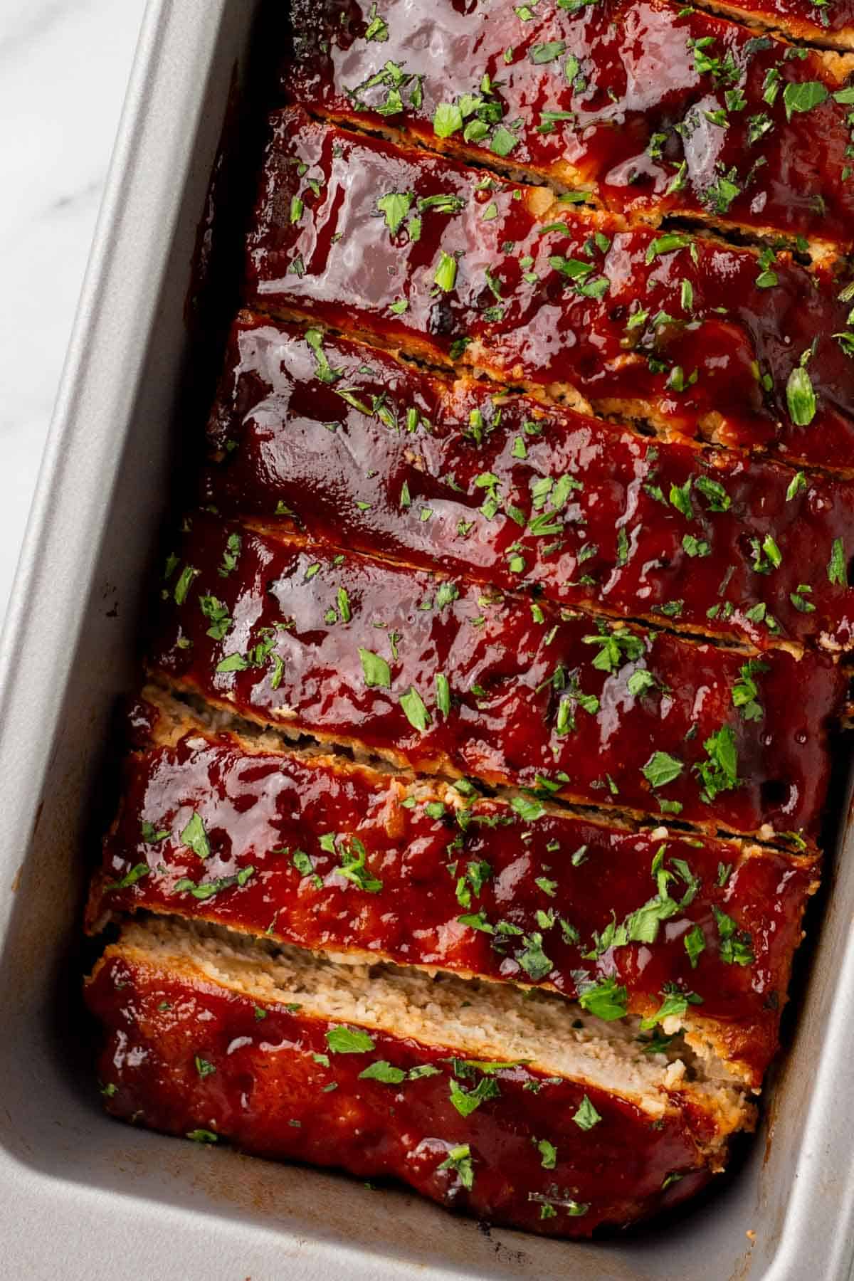Sliced turkey meatloaf topped with glaze and chopped parsley, in a baking tin.