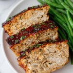 Three slices of turkey meatloaf topped with herbs are served on a plate alongside green beans.