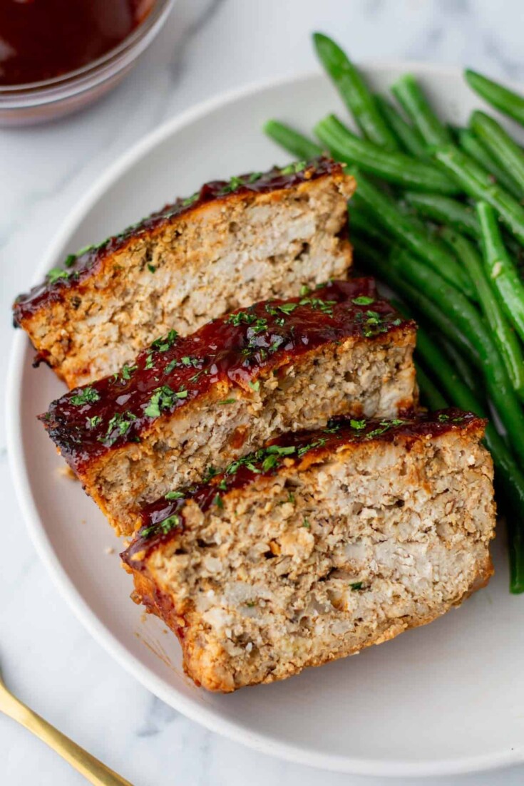 Three slices of turkey meatloaf topped with herbs are served on a plate alongside green beans.