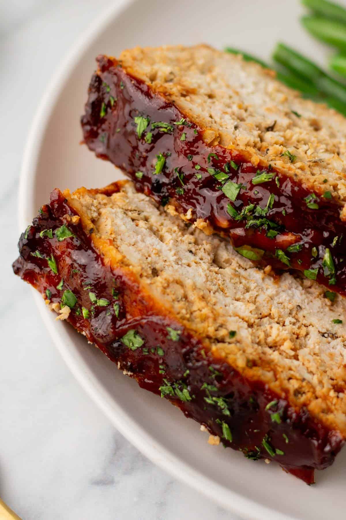 Two slices of glazed turkey meatloaf garnished with herbs on a white plate, accompanied by green beans.