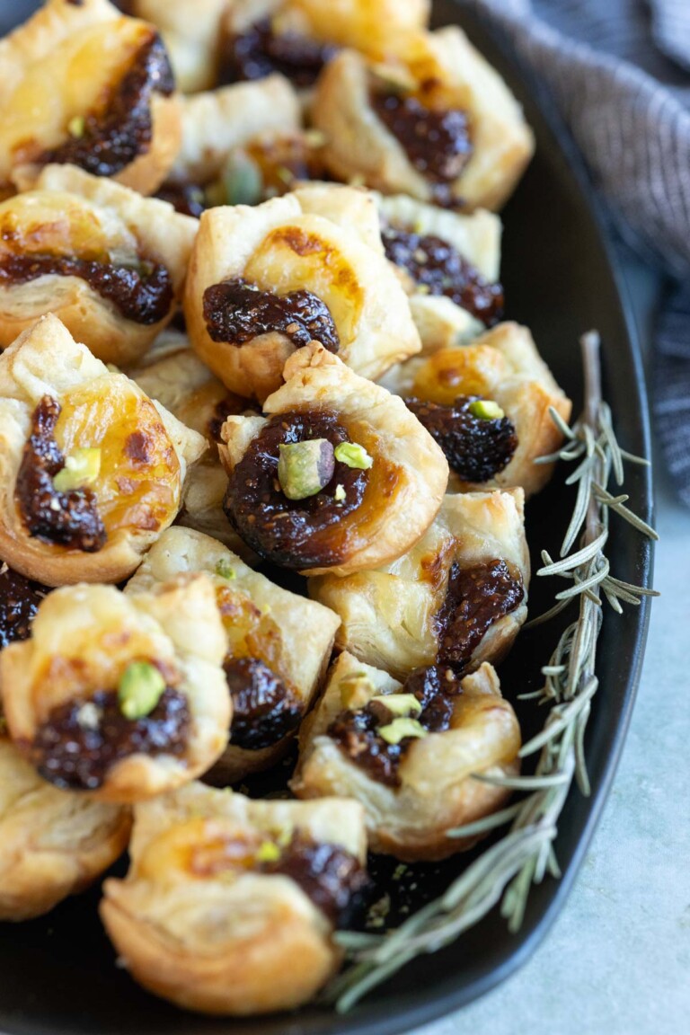 Close-up of a black platter filled with puff pastry appetizers topped with a dark jam and garnished with small green pieces, next to a sprig of rosemary.