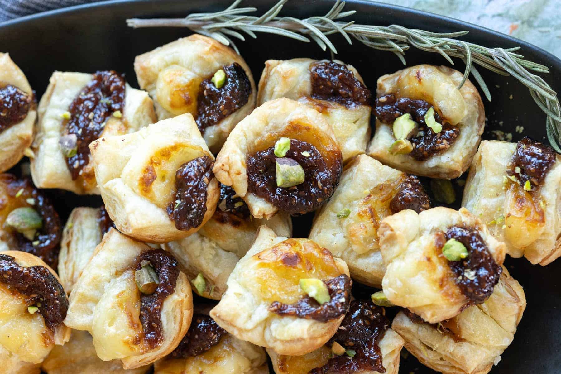 Close-up of puff pastry bites topped with fig jam, melted cheese, and garnished with pistachios, arranged on a black platter with a sprig of rosemary.