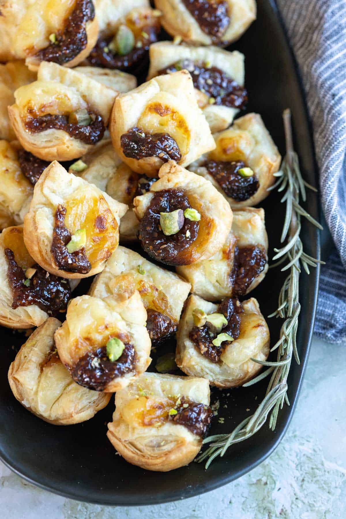 A plate of pastry puffs filled with a dark fruit spread, topped with chopped pistachios, next to sprigs of rosemary.