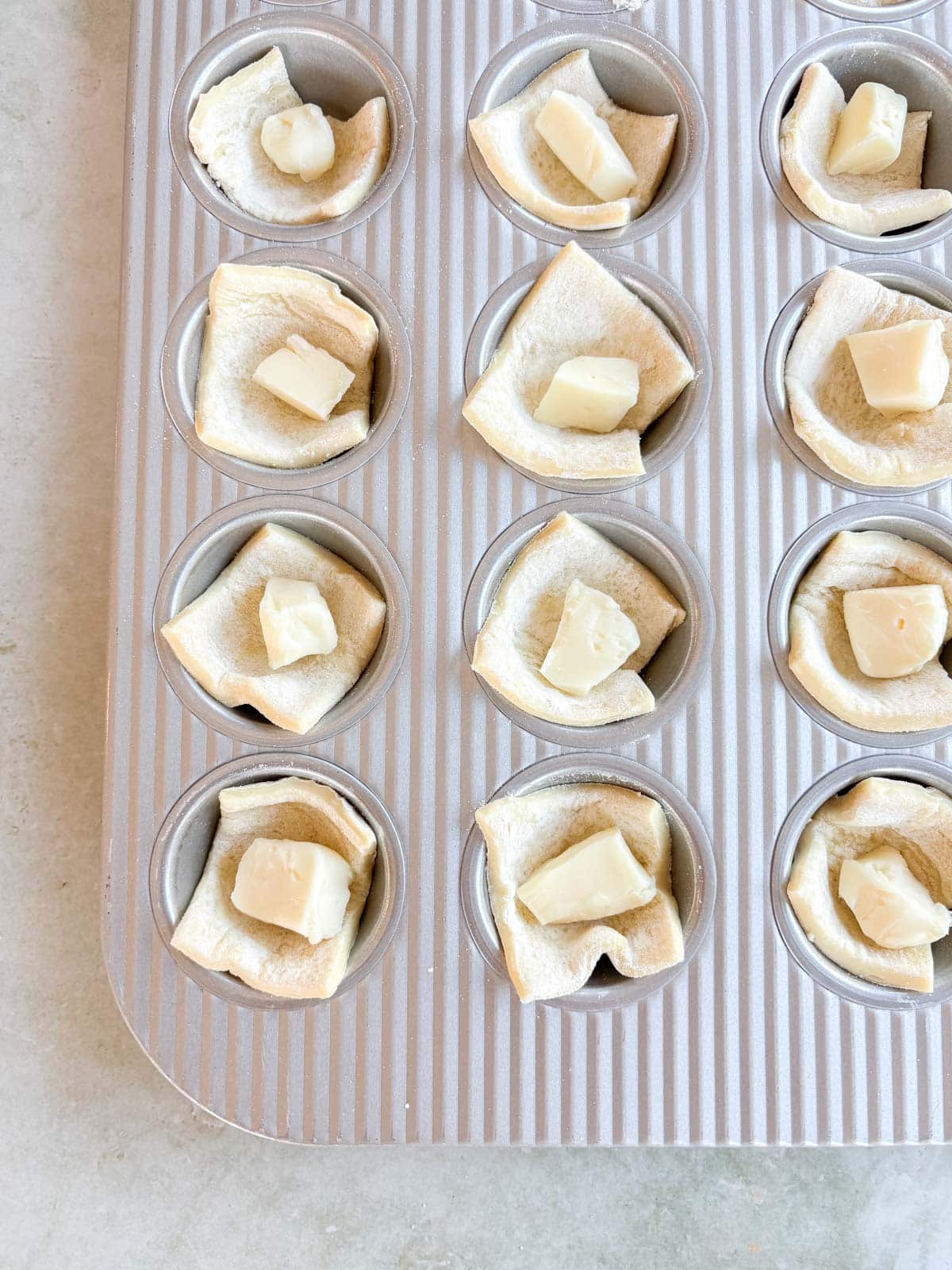 A muffin tin with puff pastry squares and cheese cubes in each cup, ready for baking.