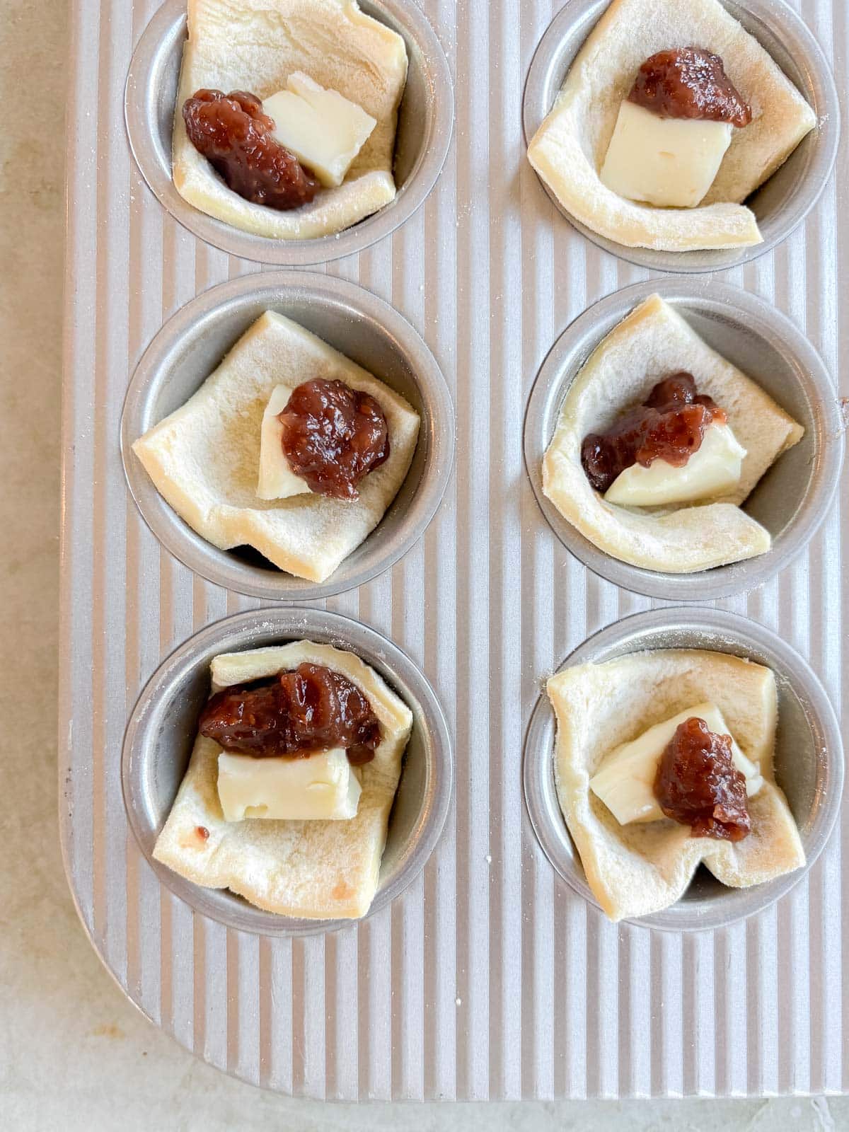 Six pastry cups in a muffin tin, each filled with a piece of cheese and a dollop of chutney.