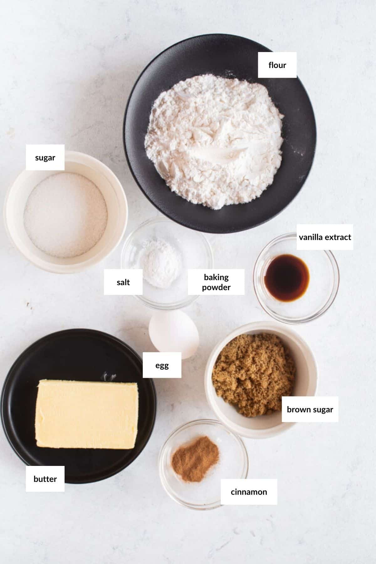 Top view of baking ingredients on a white surface: flour, sugar, brown sugar, butter, egg, cinnamon, salt, baking powder, and vanilla extract, each in separate bowls.