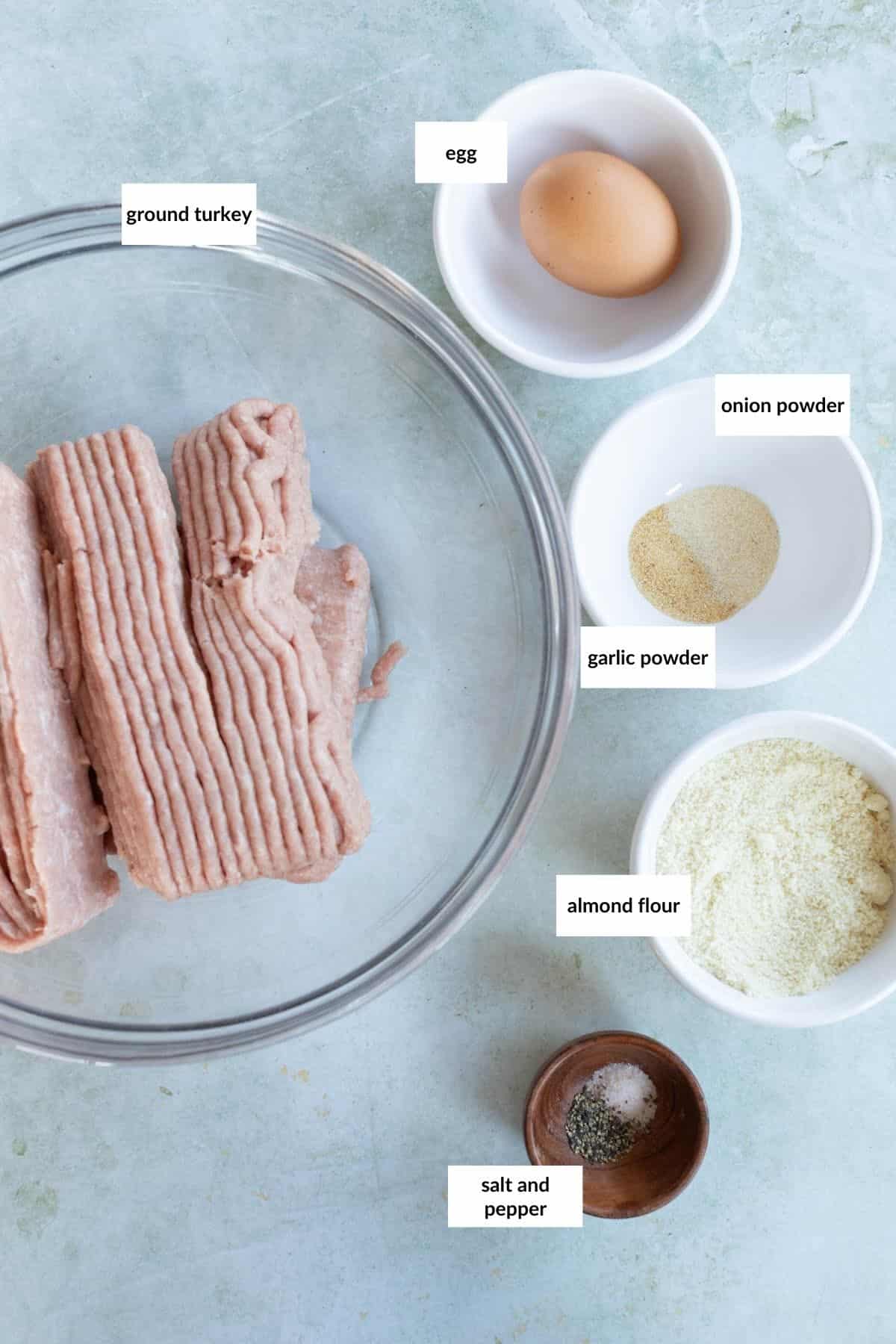 Ingredients for turkey meatballs: ground turkey, an egg, onion powder, garlic powder, almond flour, salt, and pepper arranged in bowls on a light surface.