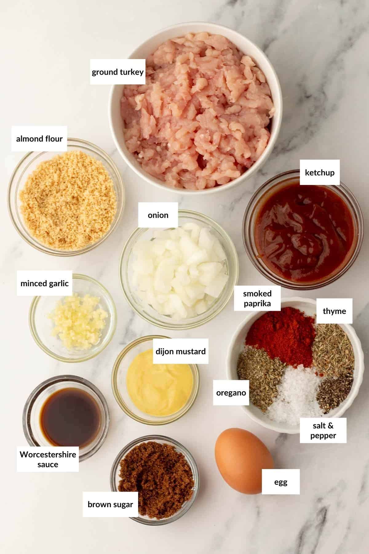 Bowl of ground turkey surrounded by bowls of almond flour, minced garlic, onion, ketchup, Dijon mustard, Worcestershire sauce, brown sugar, smoked paprika, thyme, oregano, salt, pepper, and an egg.