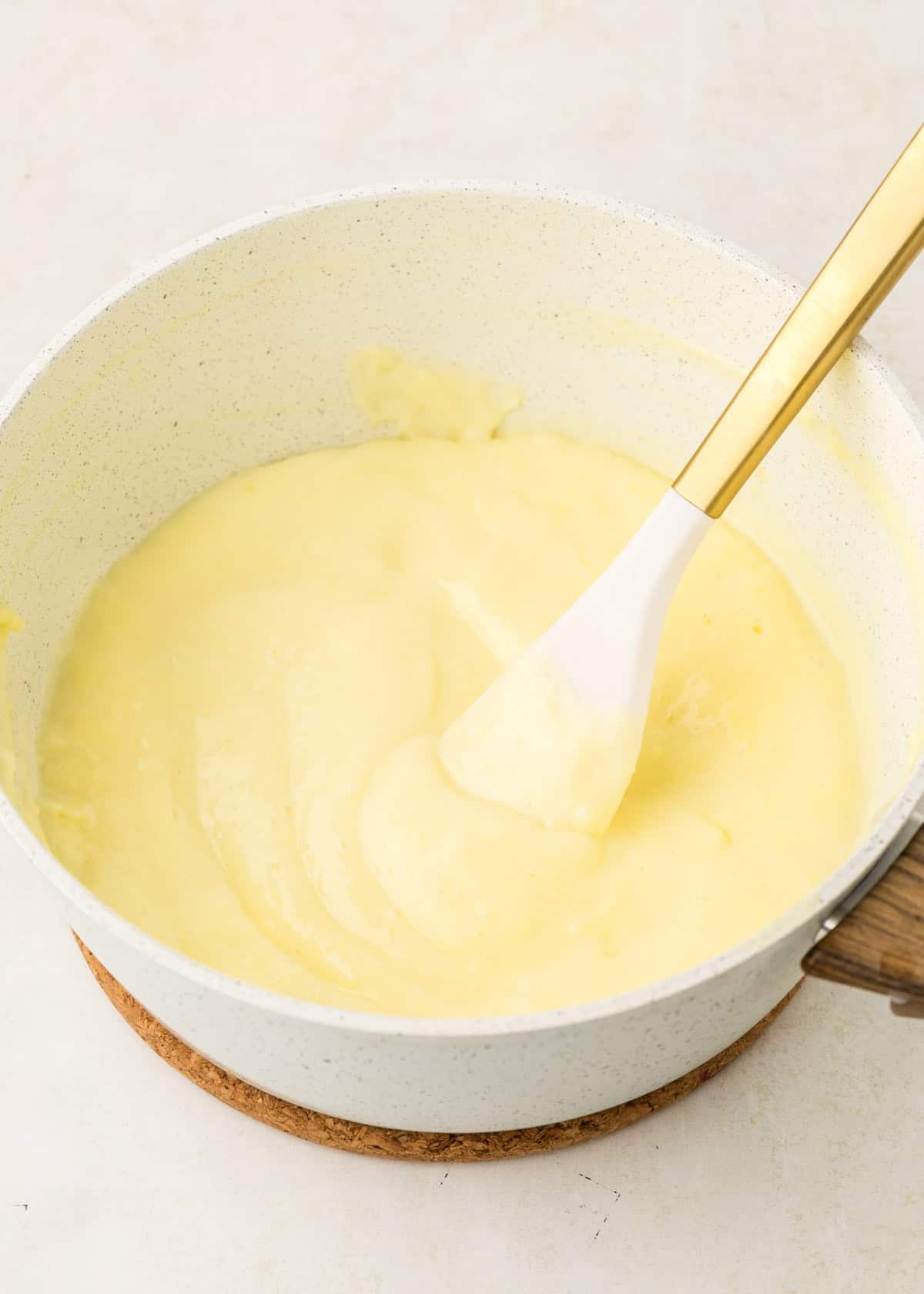 A pot of creamy yellow custard being stirred with a white spatula on a light surface.