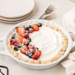 A vanilla pudding pie with whipped cream topping, garnished with strawberries, raspberries, blueberries, and powdered sugar, placed on a table next to plates and forks.
