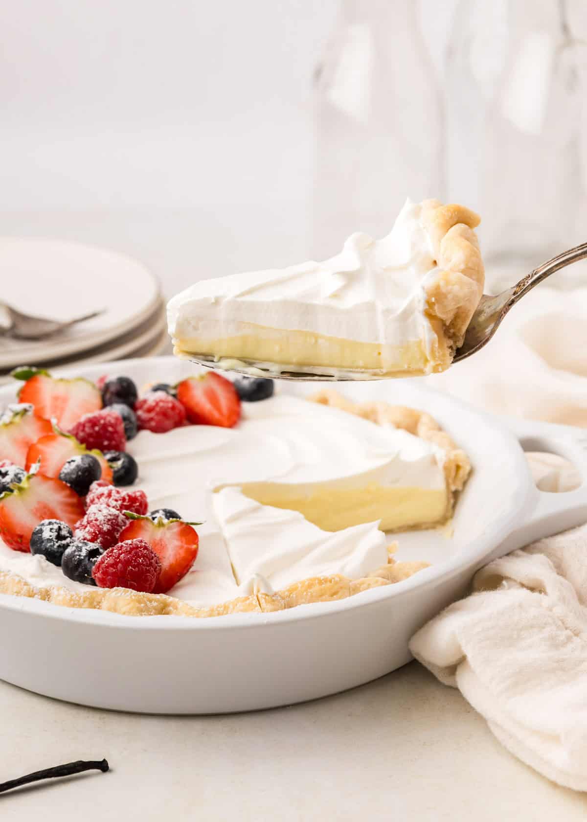 A slice of vanilla pudding pie topped with whipped cream is being lifted from a pie. The pie is decorated with strawberries, blueberries, and raspberries. Plates and bottles are in the background.