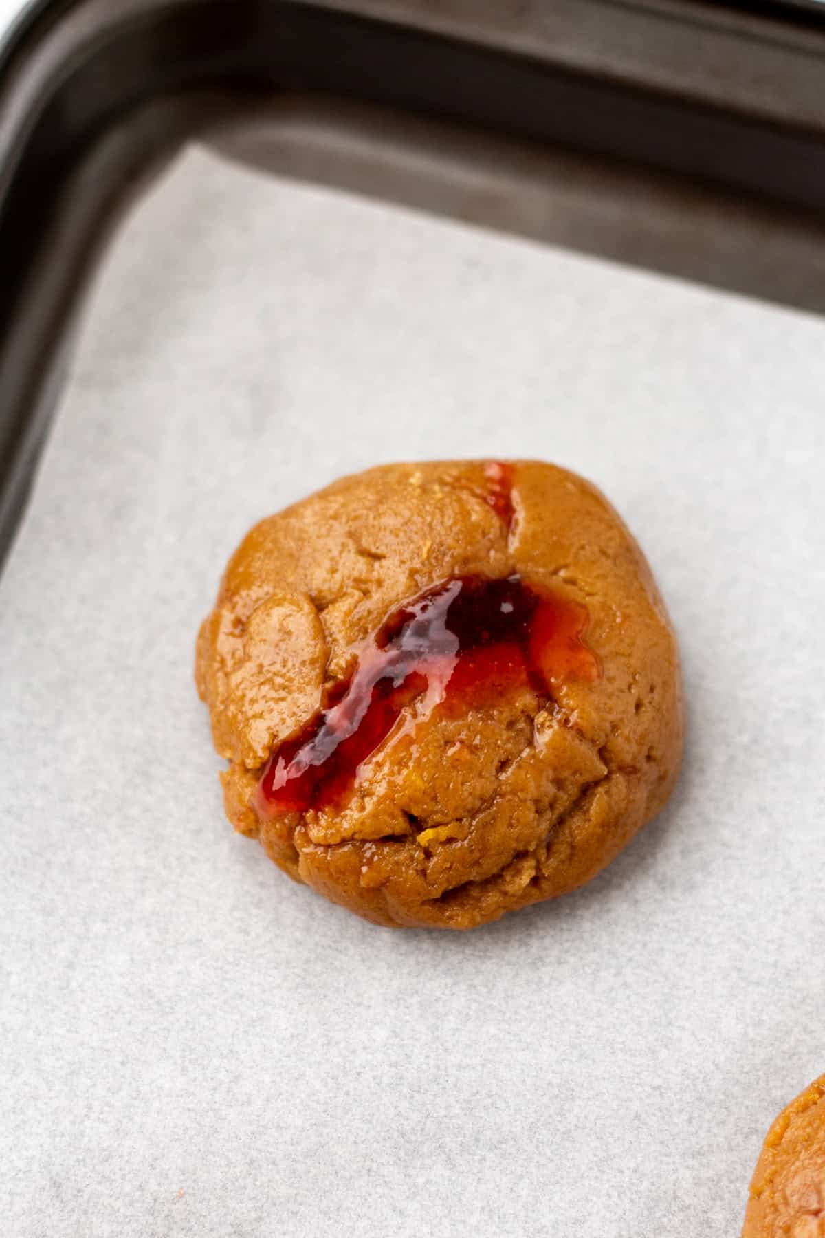 A round, brown cookie with a crack showing red jam on top, placed on white parchment paper in a baking tray.