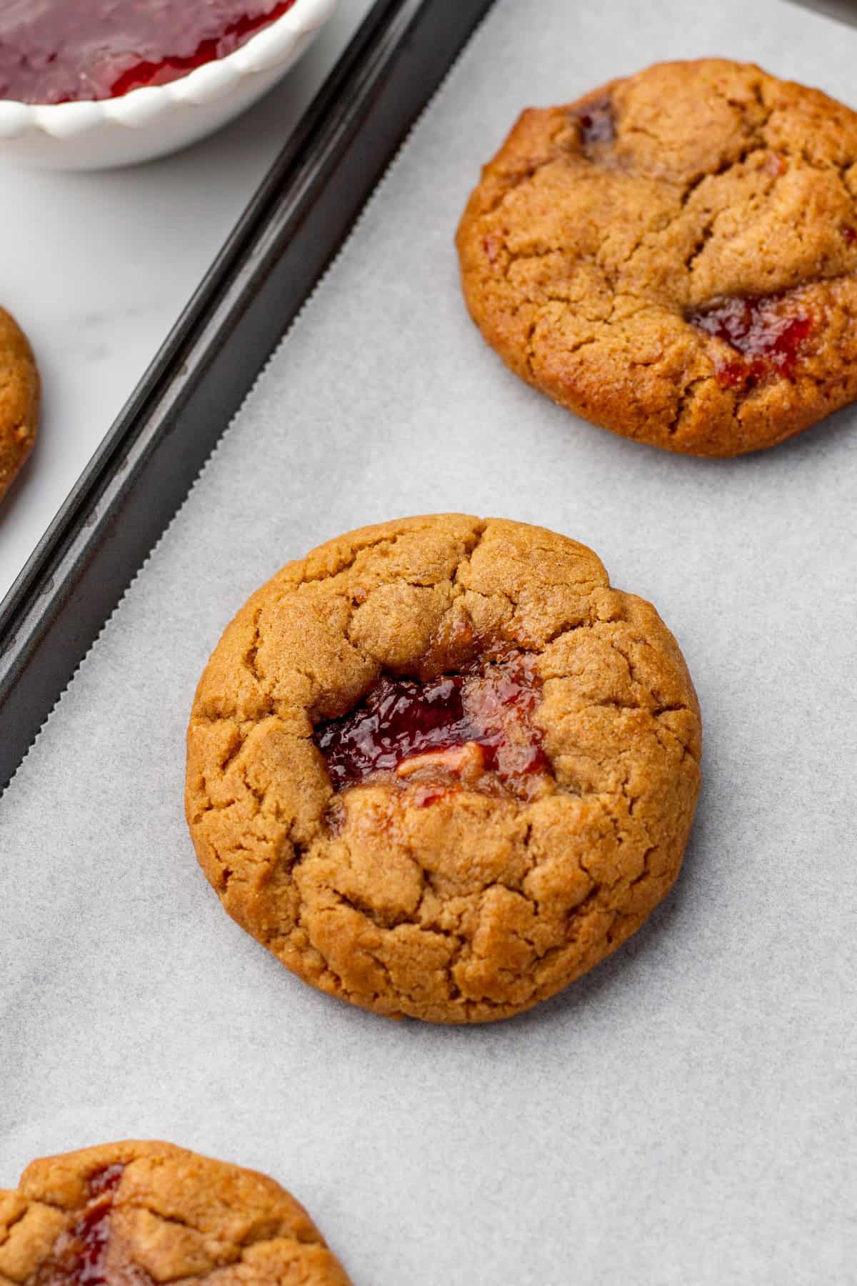 Close-up of peanut butter and jelly thumbprint cookies on parchment paper with a bowl of jelly in the background.