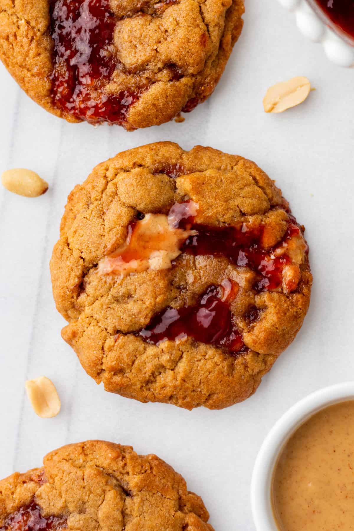 Close-up of peanut butter and jelly cookies on a white surface. Peanuts and a bowl of peanut butter are visible nearby.