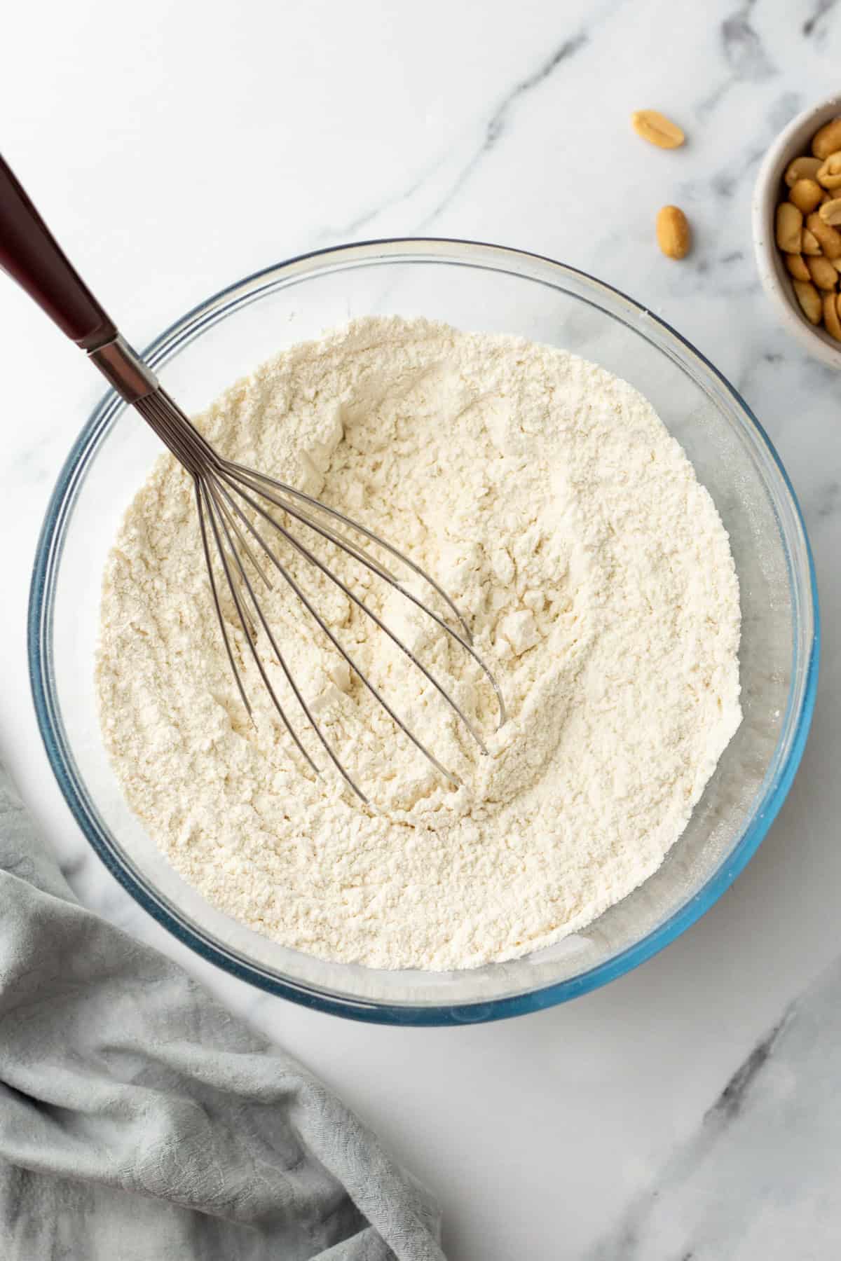 A whisk rests in a glass bowl of flour on a marble surface, with a gray cloth and a small bowl of nuts nearby.