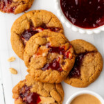 Cookies with peanut butter and jelly on parchment paper next to a bowl of jelly and a dish of peanut butter.