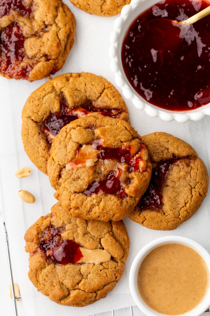 Cookies with peanut butter and jelly on parchment paper next to a bowl of jelly and a dish of peanut butter.