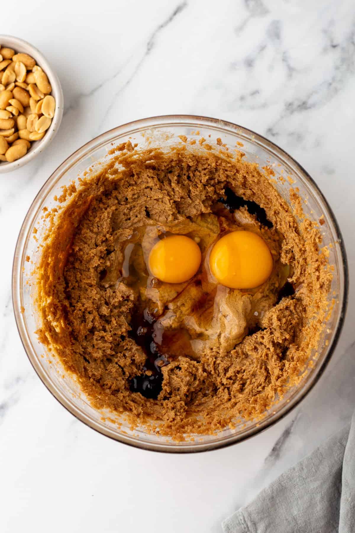 A glass bowl with brown sugar, peanut butter, two raw eggs, and vanilla extract mixed together on a marble countertop. A small bowl of peanuts is in the corner.