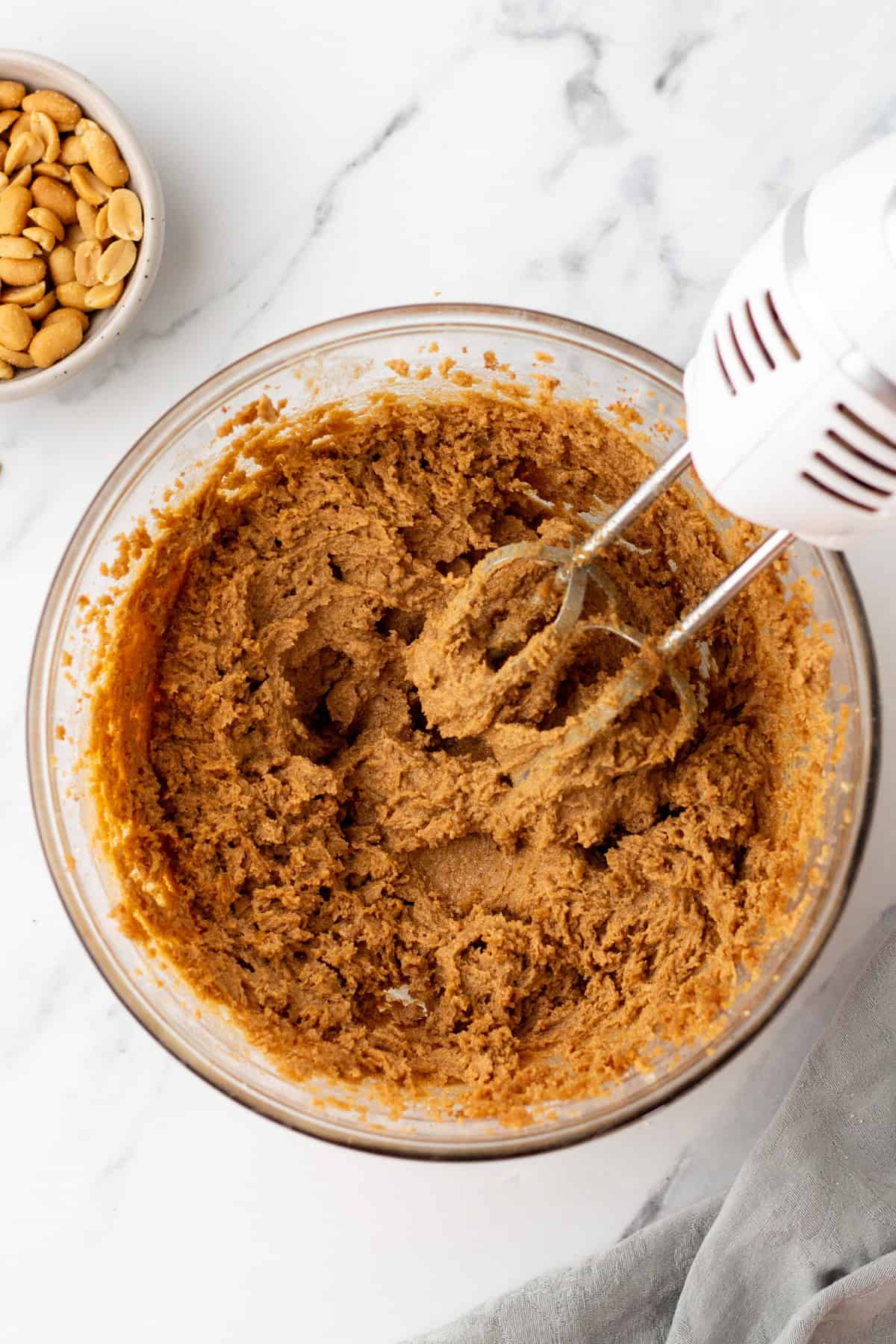 A bowl of cookie dough being mixed with an electric hand mixer, next to a small bowl of peanuts on a marble surface.