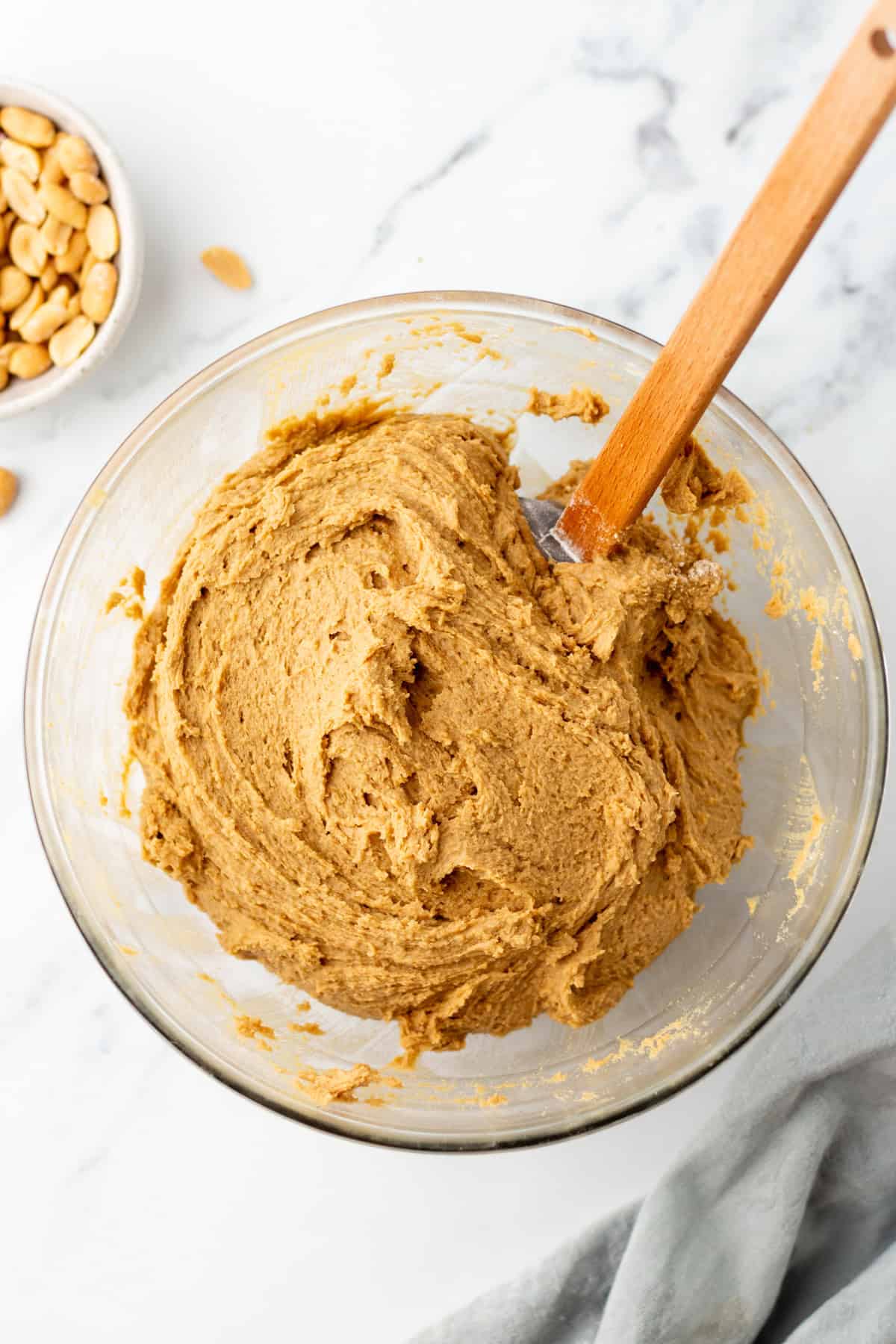 A bowl of peanut butter cookie dough with a wooden spatula, placed on a marble surface next to a small bowl of peanuts.