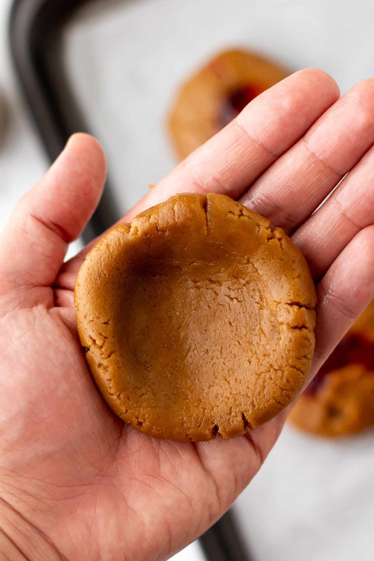 A hand holding a round, unbaked cookie dough with a thumbprint indentation.