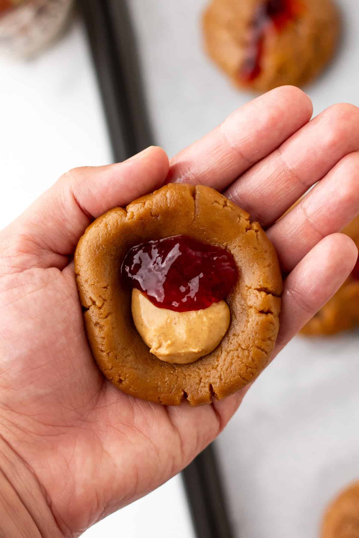 A hand holding cookie dough with a peanut butter dollop and a scoop of red jelly on top.