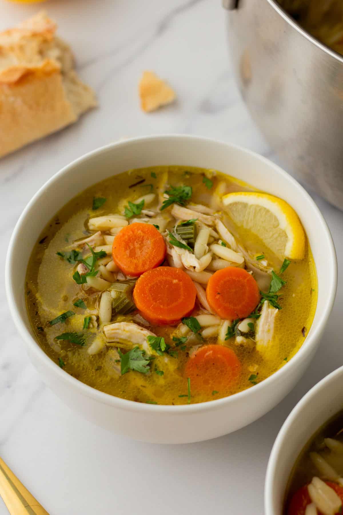 A bowl of chicken orzo soup with carrot slices, lemon wedge, and herbs, placed on a marble surface. Pieces of bread are visible in the background.