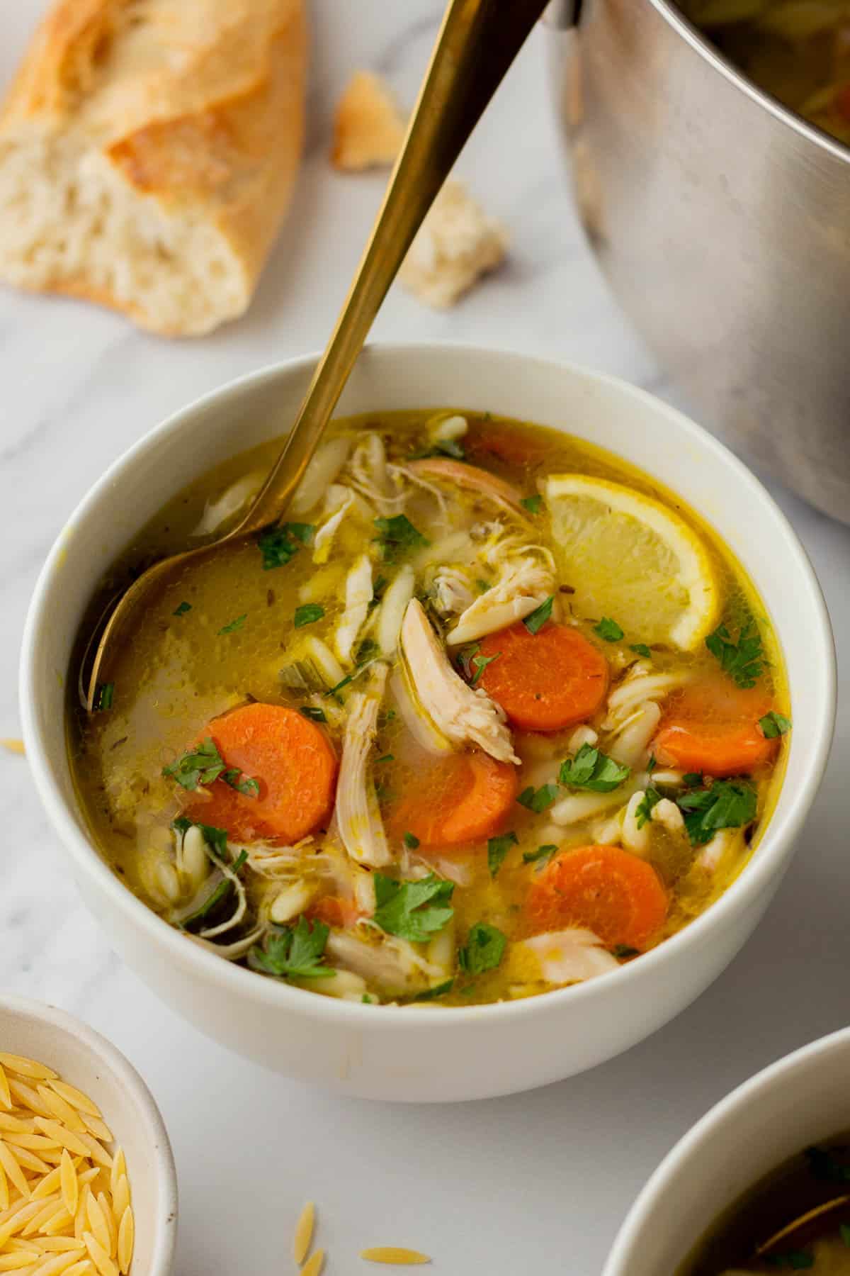 A bowl of chicken orzo soup with carrots, lemon slices, and parsley, accompanied by a spoon. Bread and a pot are partially visible in the background.