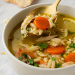 A bowl of chicken orzo soup with carrots and herbs, with a spoon lifting some soup. A piece of bread is in the background.