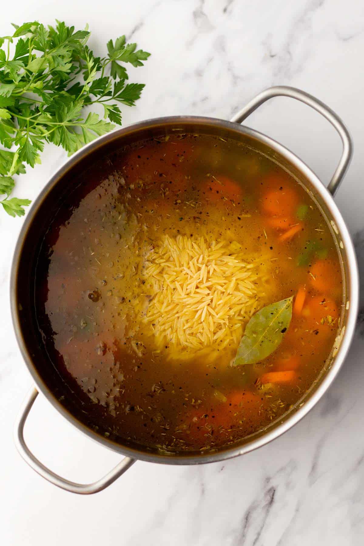A pot of soup with visible orzo, carrots, and a bay leaf on a marble surface next to fresh parsley.